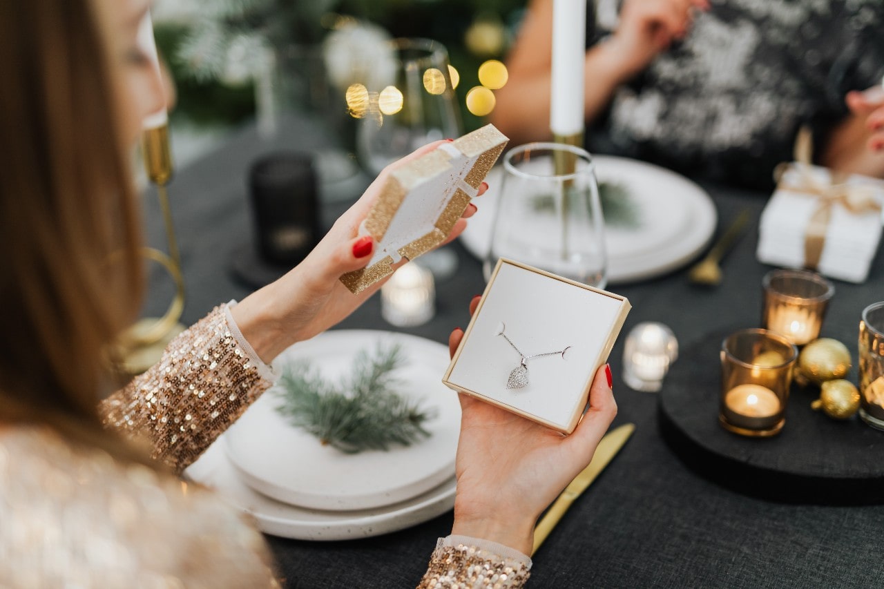 An over-the-shoulder view of a woman in a sequined dress opening a gift box holding a pendant necklace.