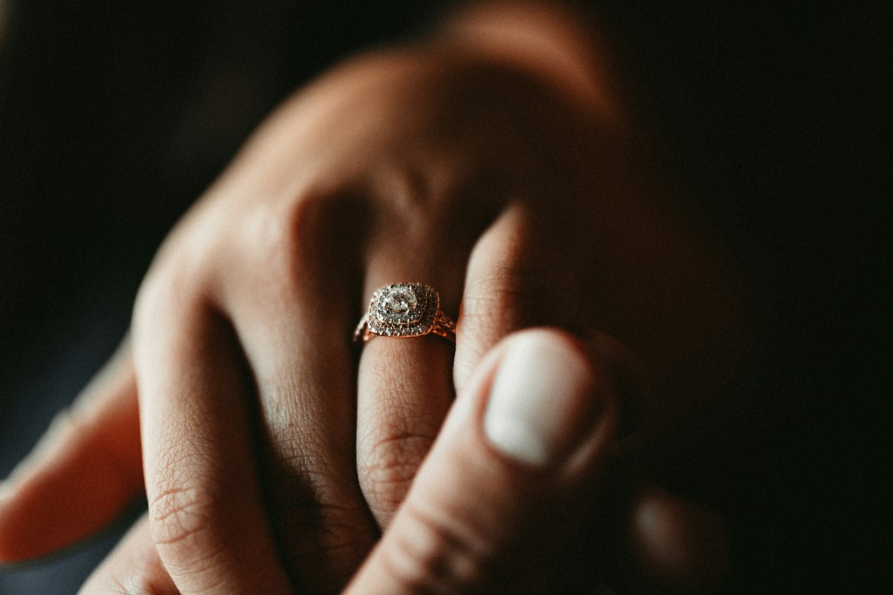 a man’s hand holding a woman’s that is adorned with a double halo engagement ring