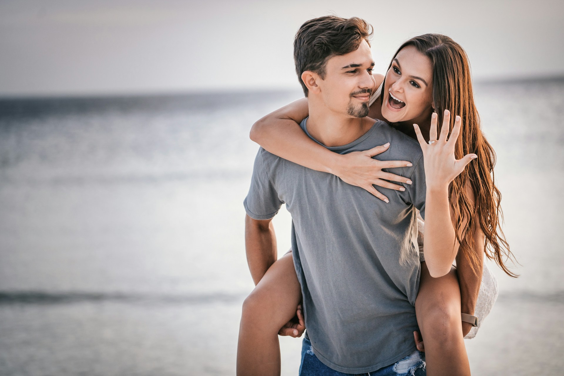 a man holding a woman on his back, the woman smiling and showing off an engagement ring