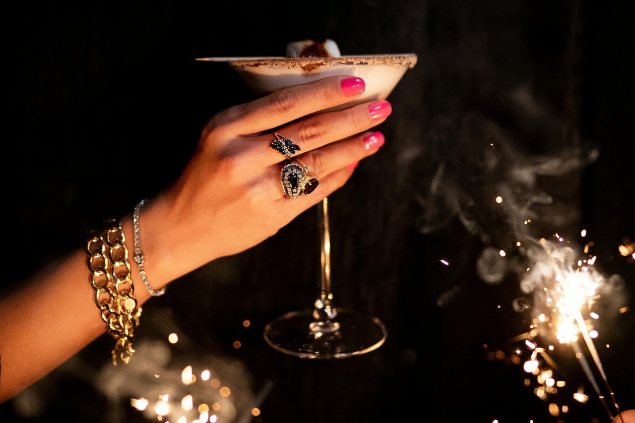 A woman’s hand holding a cocktail and wearing luxurious jewelry, surrounded by sparklers.