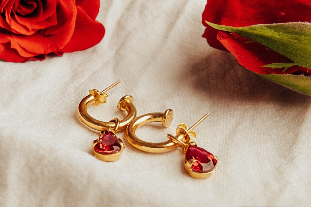 a pair of ruby drop/huggie earrings on a piece of linen surrounded by roses