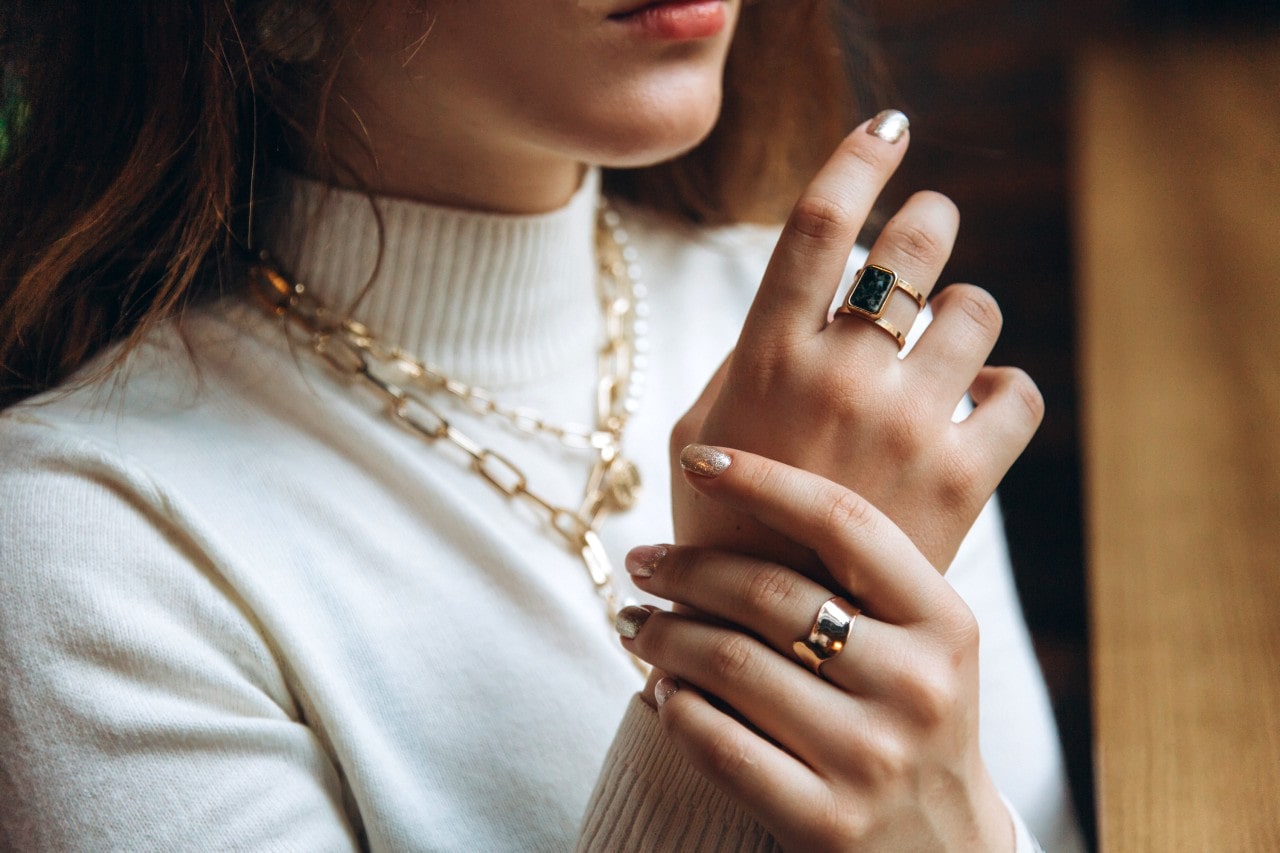 A close-up of a woman’s hands and neck adorned with chunky gold rings and layered chain necklaces over a white turtleneck. A close-up of a woman’s hands and neck adorned with chunky gold rings and layered chain necklaces over a white turtleneck.