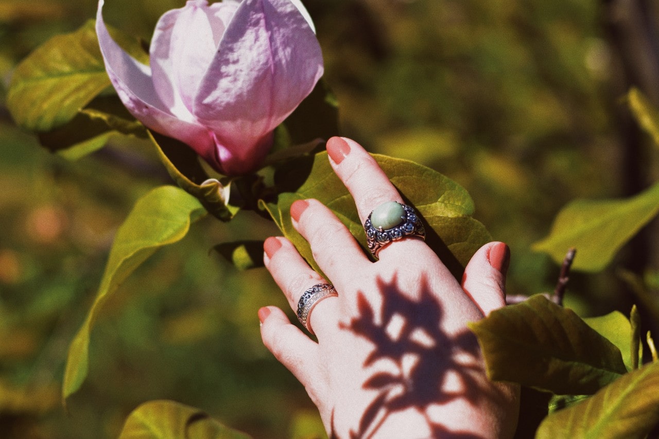 A woman reaches out for a flower, wearing a gemstone ring and two diamond rings.