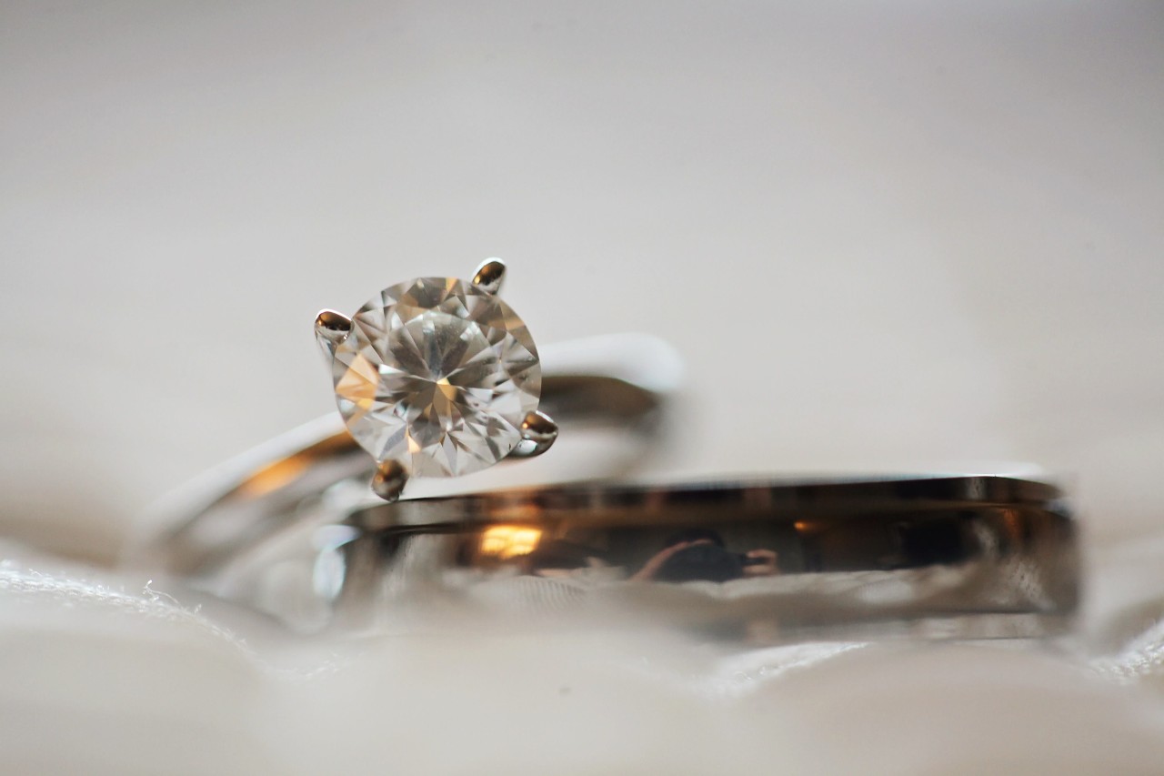 A solitaire engagement ring displayed atop a simple wedding band on a gauzy white fabric.