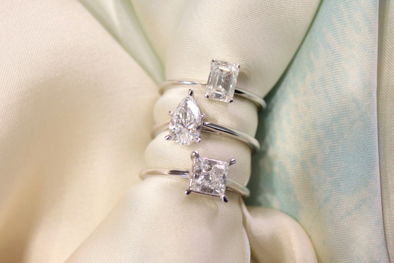 A close-up of three solitaire engagement rings of different cuts displayed on cream-colored fabric.