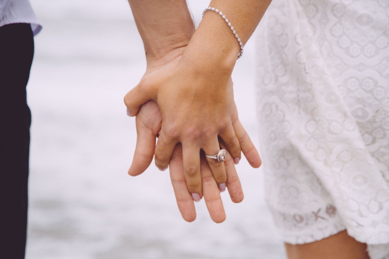 A close-up of a bride and groom holding hands, with emphasis on her engagement ring.