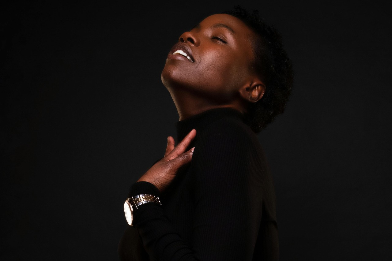 a woman wearing all black against a black background touching her neck and wearing a watch